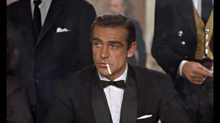 Sean Connery as James Bond in Dr. No wearing a shawl collar tuxedo with pleated shirt, white pocket square and typical sixties thin bow tie with pointed ends