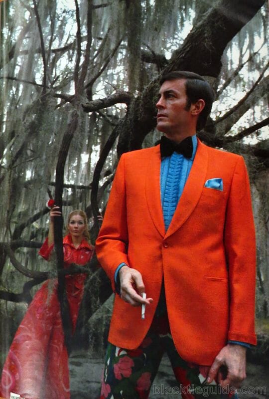 Bright orange shawl collar dinner jacket with bold blue shirt and ruffled front, matchin pocket sqaure and hib black bow tie and bold flowery pants