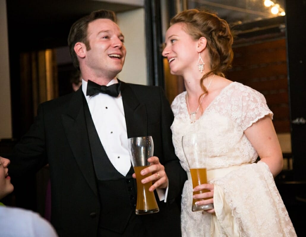 Black Tuxedo with deep v-cut evening vest, white shirt with 3 shirt studs, black bow tie next to the bride, both drinking wheat beer.