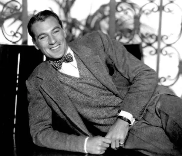 Gary Cooper wearing a herring bone suit and grey v-neck sweater and bow tie