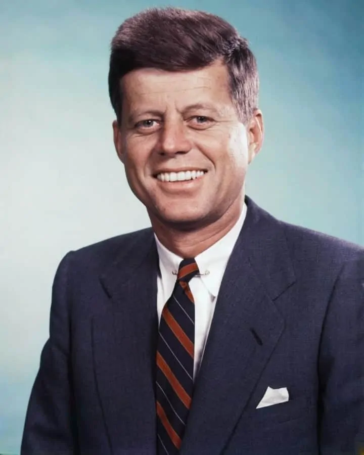 John F Kennedy smiling in this photo in a navy jacket paired with a stripe tie and white linen pocket square