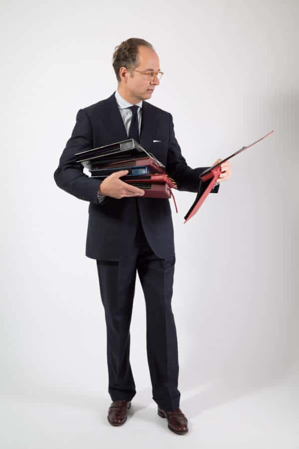 Bernhard Roetzel wearing burgundy loafers with grey windowpane suit, dark knit tie, stripes shirt and fabric books