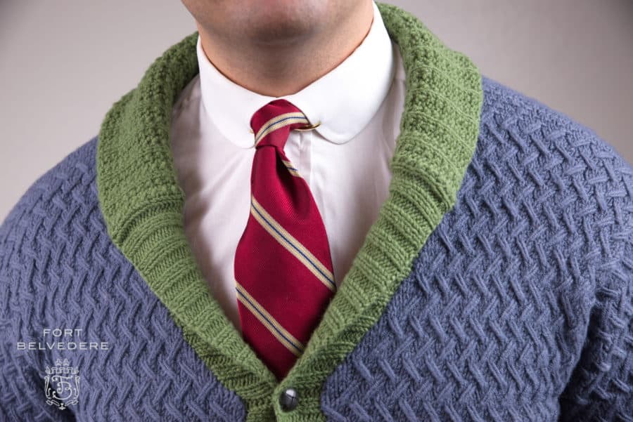 A white shirt with a club collar, worn with a collar pin and red striped tie, under a blue and green cardigan sweater.