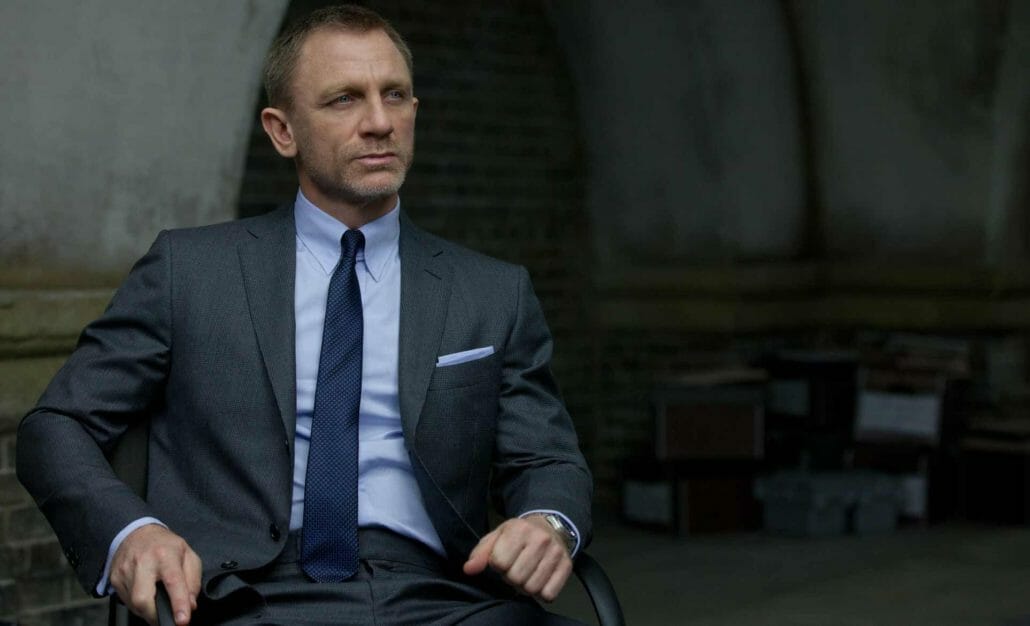 Bond wearing a dark gray suit, light blue shirt, pocket square, and navy tie with polka dots.