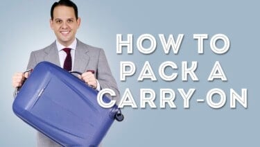 Raphael wearing a 2-piece gray suit, burgundy knit tie and pocket square and is holding a blue carry-on suitcase