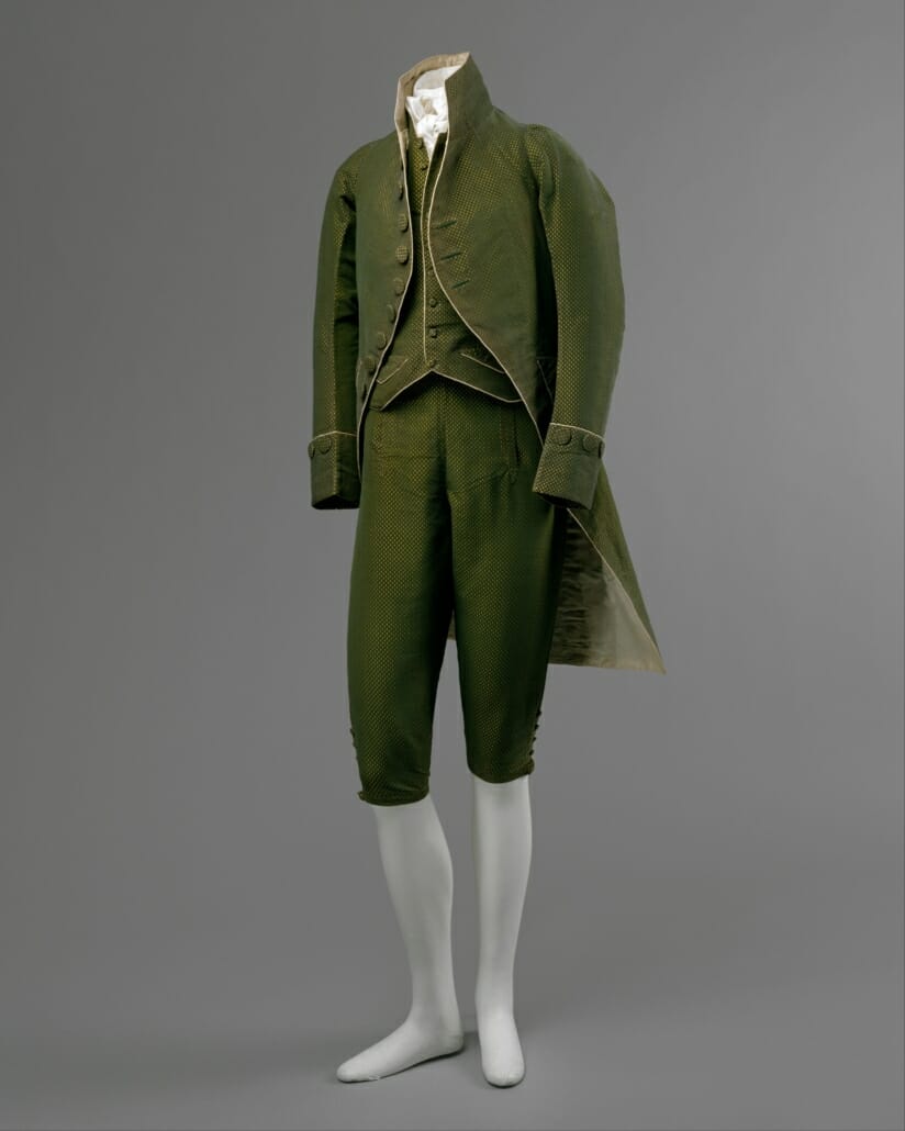 This outfit from c. 1790 features a coat with narrow sleeves and a cut-away skirt, a short vest, and a pair of breeches. Some opulence remains in its luxurious fabric.