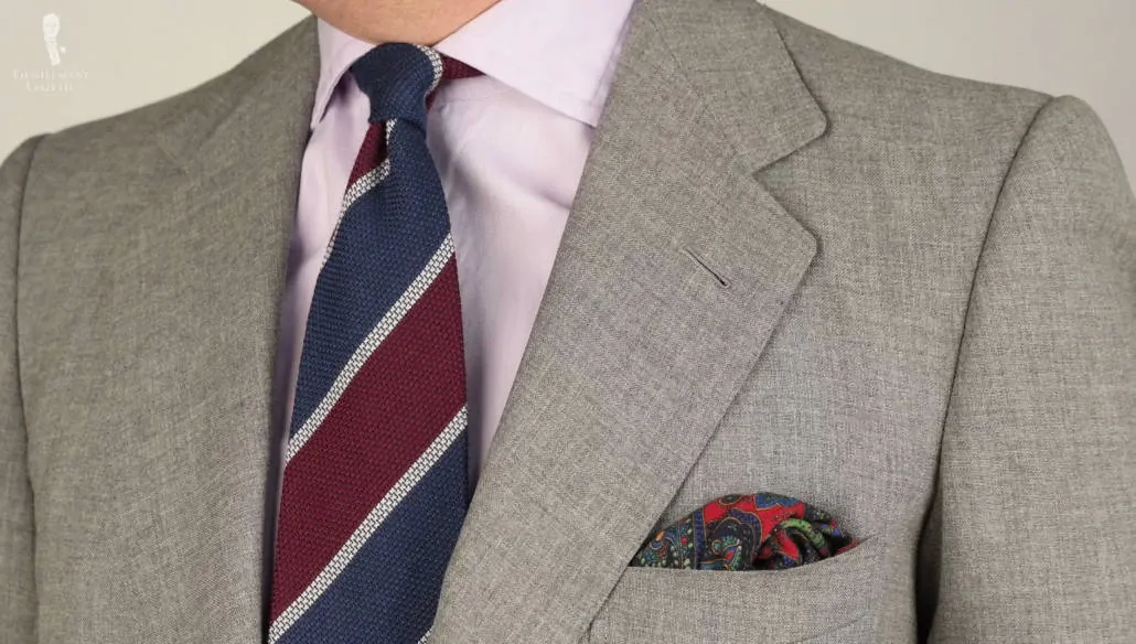 A Cashmere-Wool Grenadine Tie in Dark Blue, Burgundy, Light Grey Stripe from Fort Belvedere, paired with a light gray suit, lavender shirt, and patterned pocket square