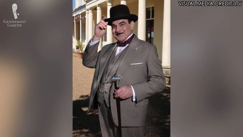 Hercule Poirot in brown three piece stripe suit and a hat, and carrying a cane