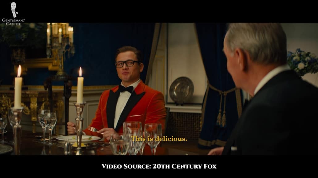 Eggsy in an orange velvet dinner jacket with black velvet peak lapels and side vents, a white shirt with regular buttons, a black velvet batwing bow tie, white TV-folded pocket square, black trousers, and gold cufflinks.