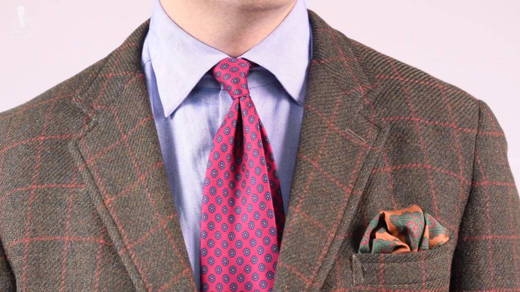 Raphael wearing a green tweed jacket with an orange windowpane, bronze yellow pocket square, blue shirt, and a madder silk tie in ruby red.