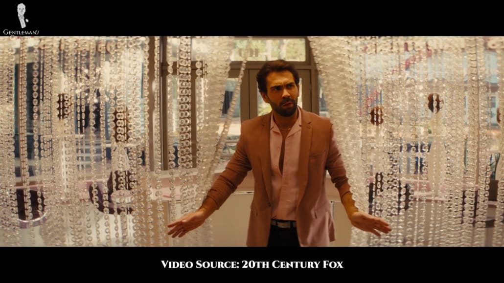 The henchman Angel wearing a mauve jacket with narrow lapels, a pink shirt with a Cuban collar, a gold necklace, dark jeans, and a white belt.