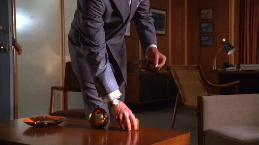 Don Draper sporting a French cuff and gunmetal cufflinks. He's also donning a pin-striped gray business suit. 