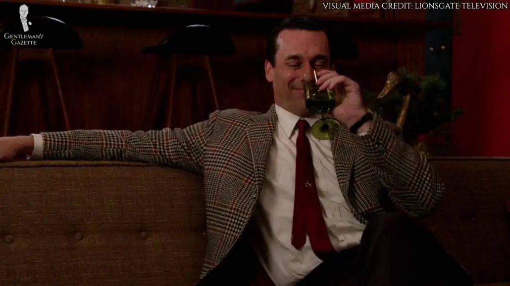 Don Draper sitting with a glass of wine in his hand. He is currently wearing a houndstooth sport coat, dark red slim tie, and a white dress shirt with pointed collars. 