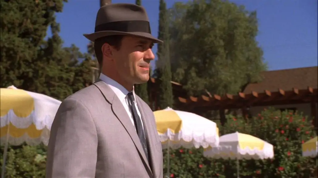 Don Draper standing by the pool side wearing a gray fedora hat with black band, gray suit, and white dress shirt. 