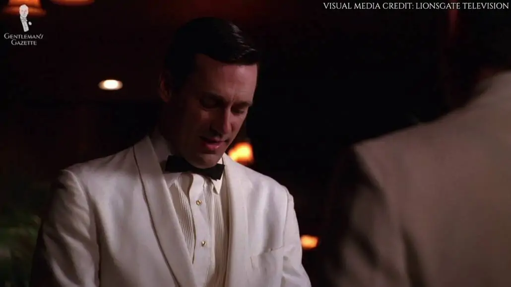 Don Draper donning a single button white dinner jacket with a very narrow shawl collar with a flat pocket.He's also wearing a white pleated shirt with gold shirt studs and a slim batwing bow tie. 
