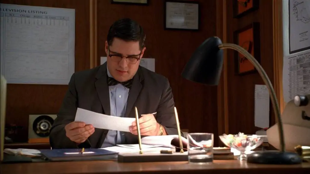 Harry Crane sitting at his desk and looking at a letter. He is wearing an unbuttoned suit jacket, a patterned bow tie, and a dress shirt