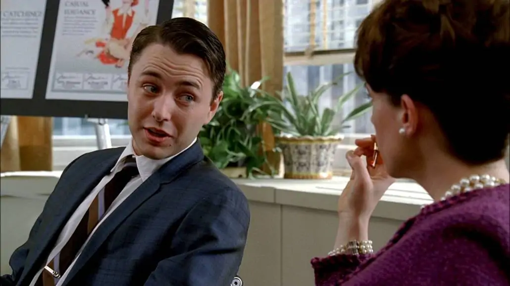 Pete Campbell having a conversation with a colleague; he is wearing a lighter than navy suit and a striped tie.