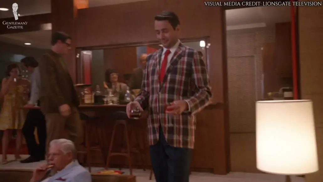 Pete Campbell walking across the room with drinks in both his hands. He's wearing a madras jacket, orange tie, and navy trousers.