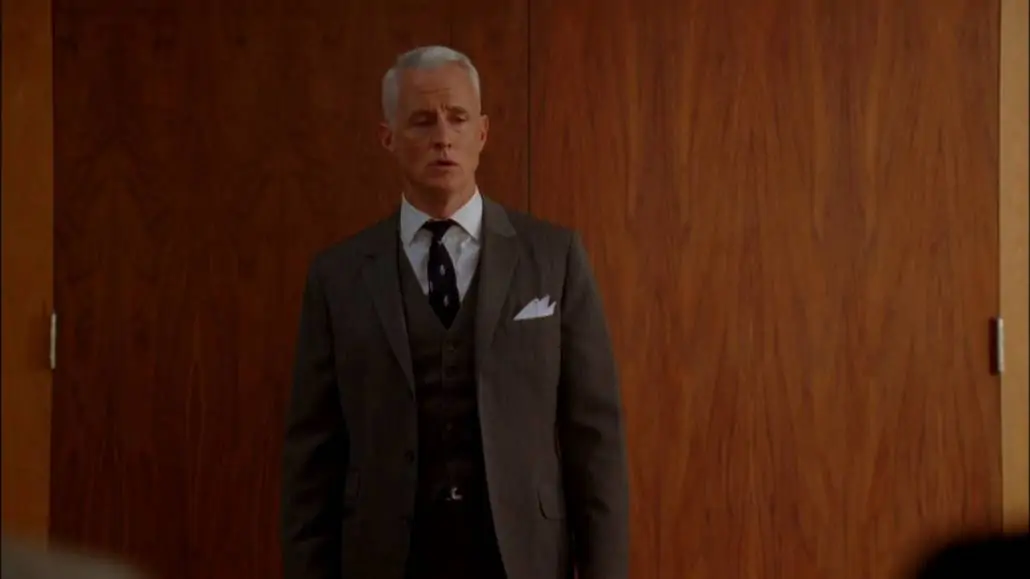 Roger Sterling wearing a three-piece charcoal suit, white pocket square, white dress shirt, and a navy tie. 