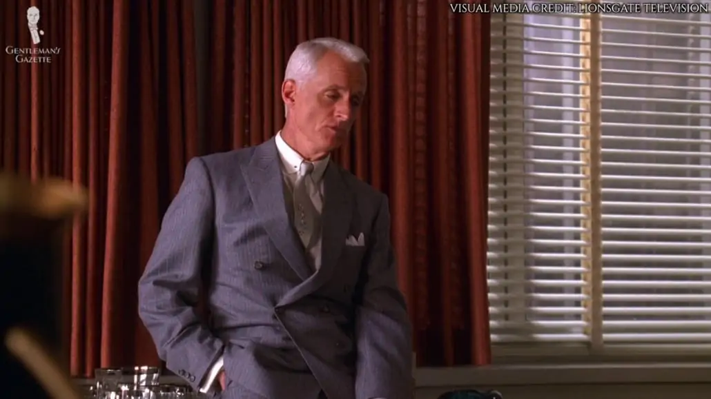 Roger Sterling wearing a slate blue, double-breasted suit with fine stripes in pale gray and orange with the bottom button undone. 