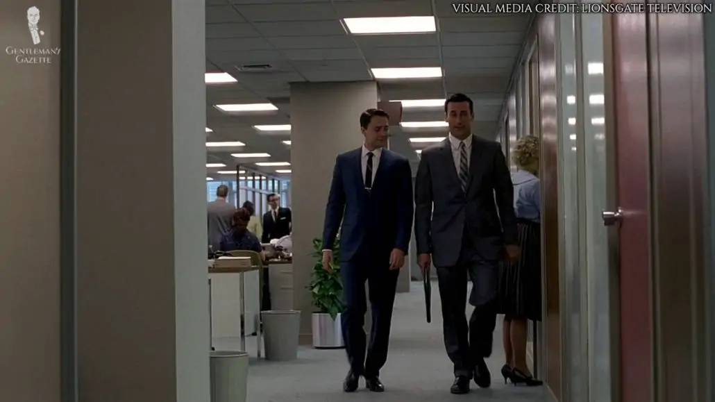 Don Draper and Peter Campbell walking along the hallways. Both are wearing business suits. 