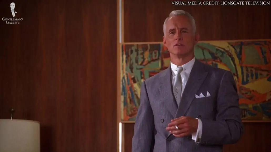 Roger Sterling wearing a slate blue, double-breasted suit with fine stripes in pale gray and orange, silver collar bars, silver, palish-blue tie with a fairy dust medallion detailing, white dress shirt with French cuffs, and silver cufflinks. He is also holding a cigarette in his left hand.