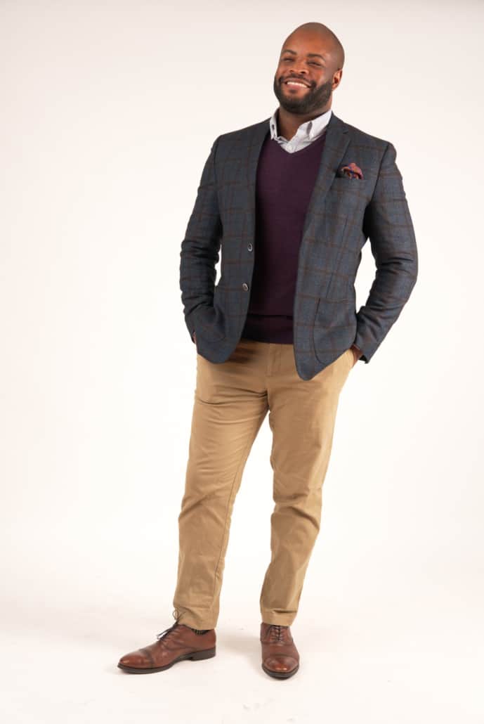Kyle wearing a gray checked unstructured sport jacket with a pair of khaki chino pants, a dress shirt, a purple sweater, and brown dress shoes