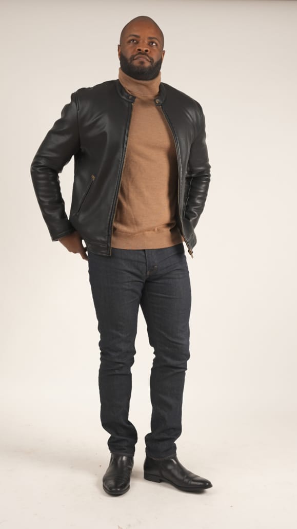Kyle wearing a black motorcycle leather jacket, tan turtleneck sweatshirt, denim jeans, and black boots.