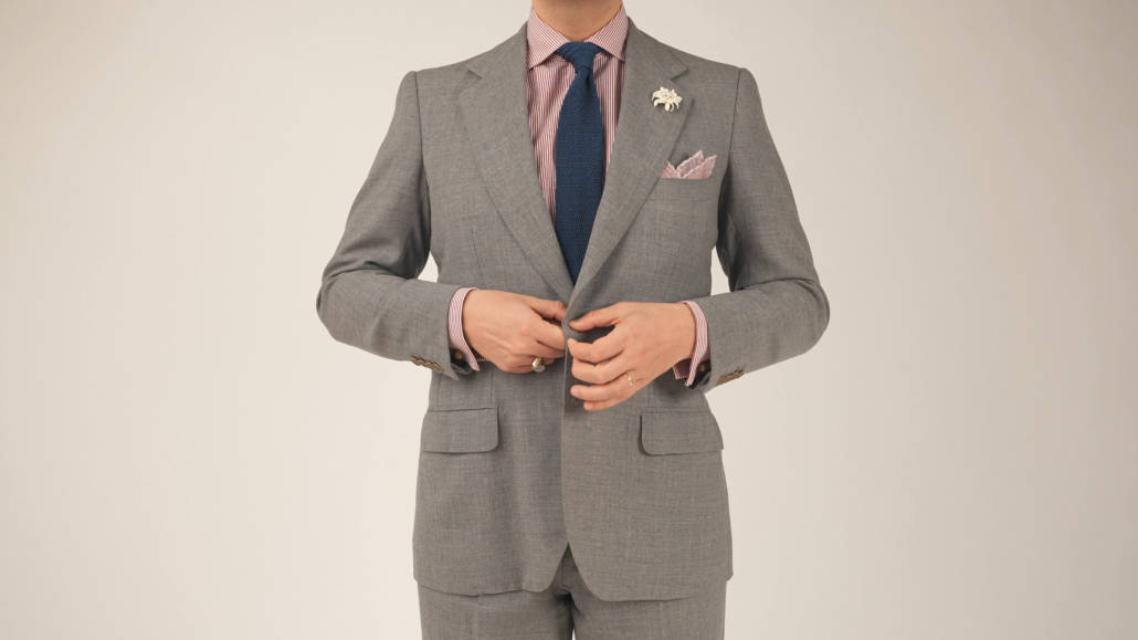 Screen A7S 167000 06 Raphael buttoning his suit jacket; he's wearing a gray suit with flap pockets, Light red striped dress shirt, navy tie, white boutonniere, and a light red pocket square from Fort Belvedere.