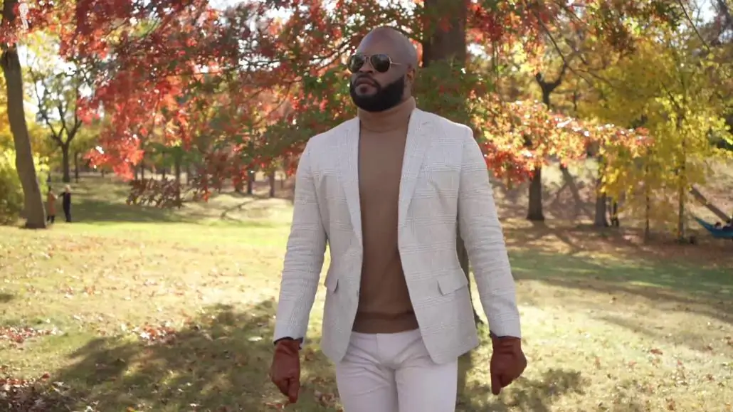 Kyle outdoors wearing an off-white checked unstructured jacket, tan turtleneck sweater, white chinos and aviator sunglasses.