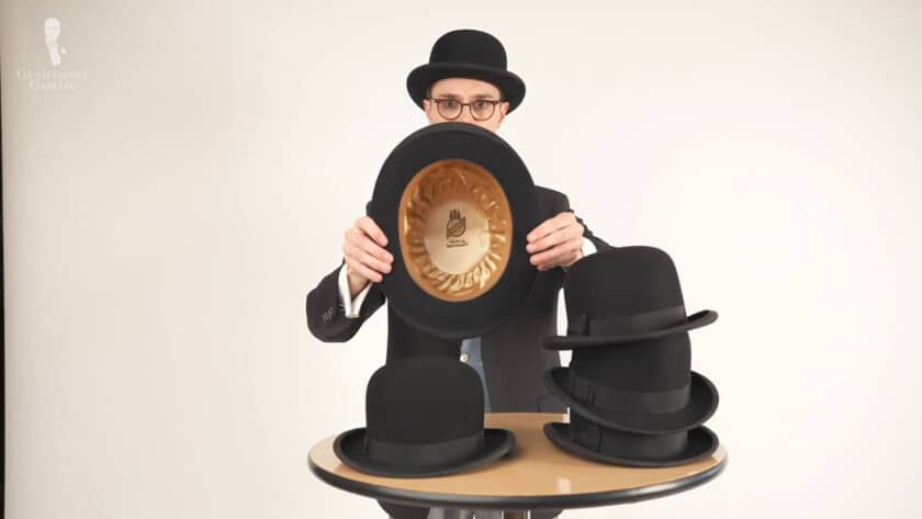Preston holding showing a bowler hat's crown. There are also a stack of other bowler hats on top of the table.