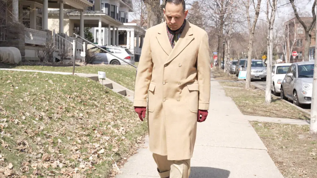 Raphael walking outdoors in a tan camel hair coat paired with suede burgundy gloves and burgundy scarf.