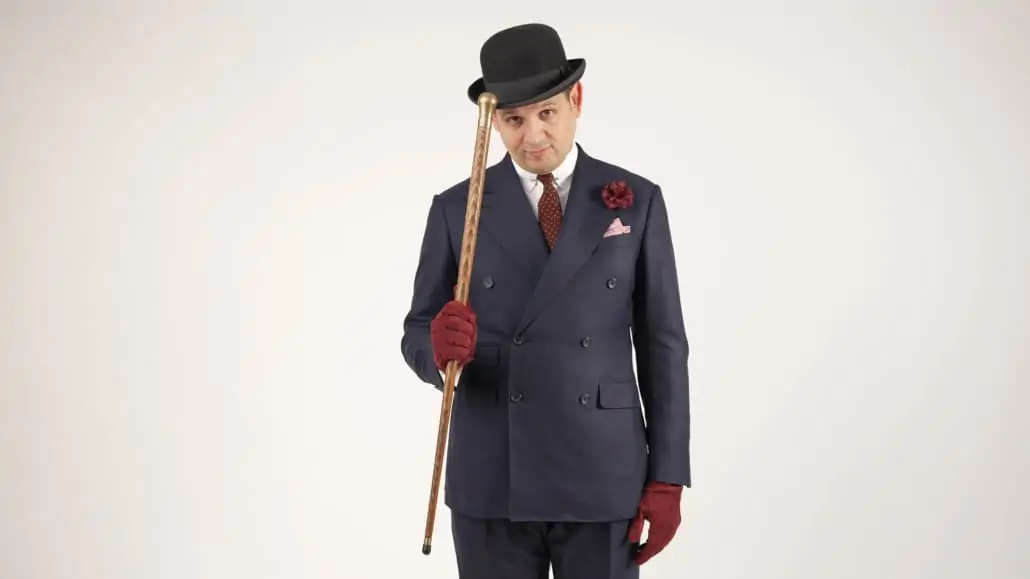 Raphael wearing a navy suit, black bowler hat, and accessories with a touch or red/burgundy. He's also holding a walking cane.