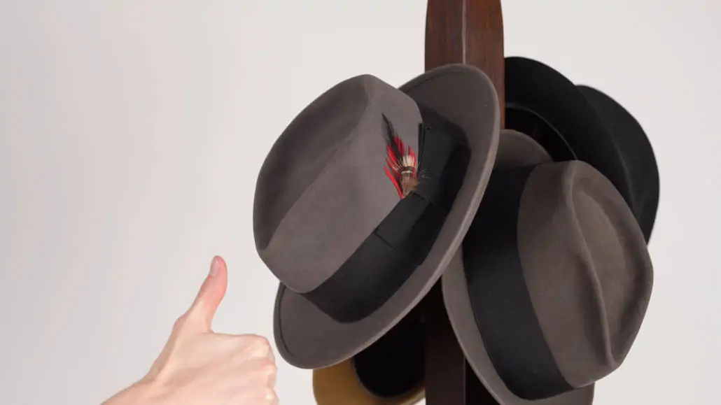 Preston giving a thumbs up sign next to a hat rack with fedoras. 