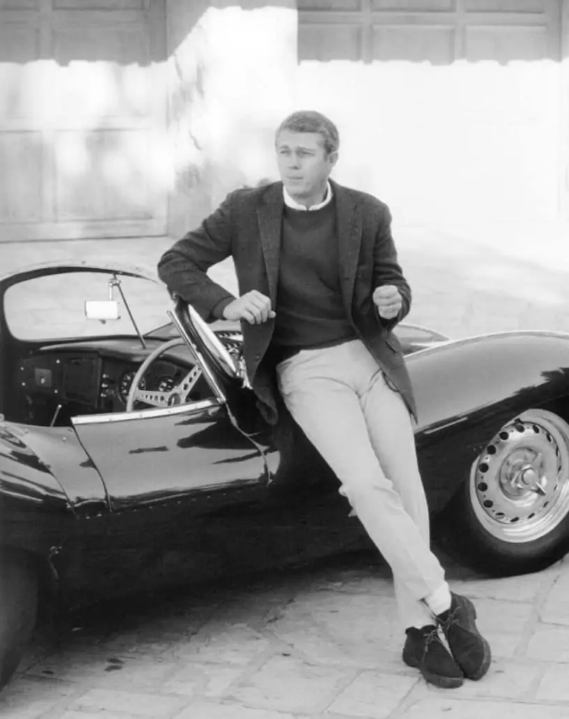 Steve McQueen wearing a pair of khaki pants with some white socks, chukka boots, crewneck shirt, sweater and a coat.