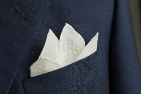 A white pocket square in a suit pocket, embroidered with an initial 