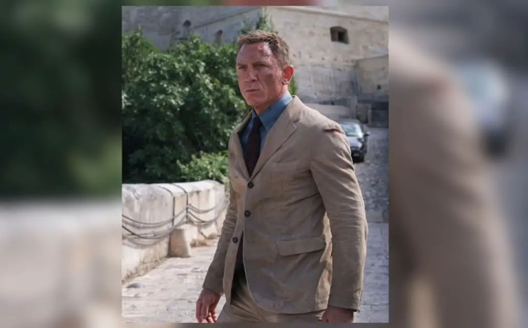 Daniel Craig as James Bond in No Time To Die, wearing a sand corduroy unstructured suit, buttondown cornflower blue shirt, and a maroon silk tie with a repeating blue tile pattern