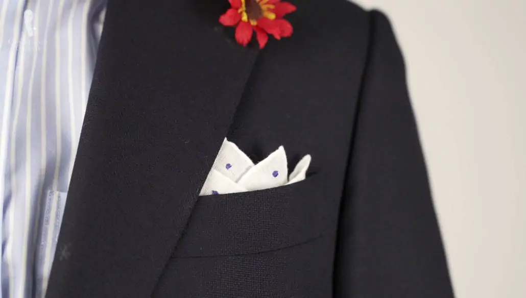 Navy suit pared with blue and white shirt accessorized with white pocket square with blue polka dots and red boutonniere.