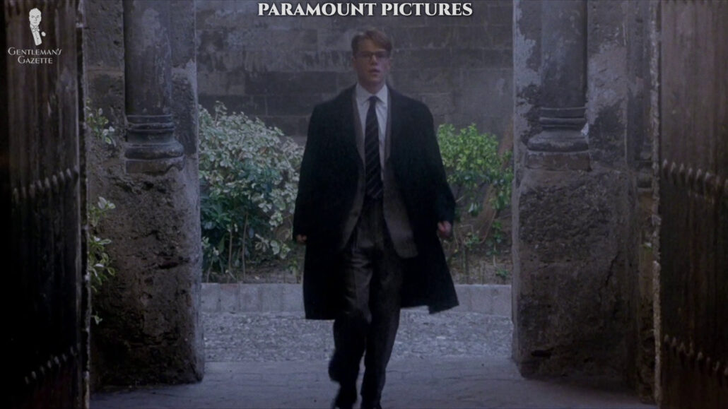 Tom Ripley wearing a glen check suit with a white shirt striped tie and a dark overcoat.