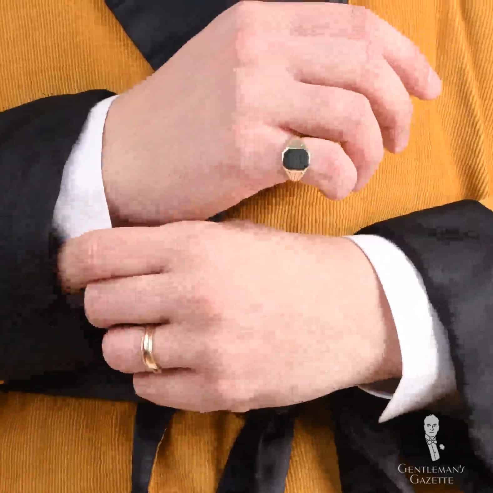 A photograph of a pair of hands with a gold wedding ring on the left ring finger and a pinky ring on the right hand