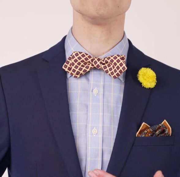 A photograph of Preston wearing a dark navy suit with a printed bow tie, a pocket square with a tinge of gold and a yellow dandelion boutonniere.