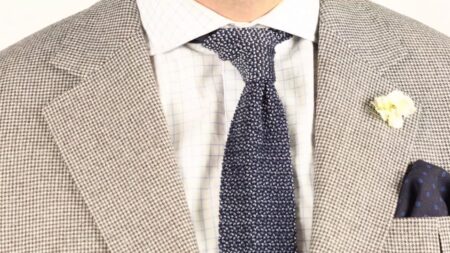 A navy blue wool challis pocket square with blue polka dots paired with a houndstooth jacket, a checked shirt, a white carnation boutonniere, and a navy and light blue two-tone knit tie.