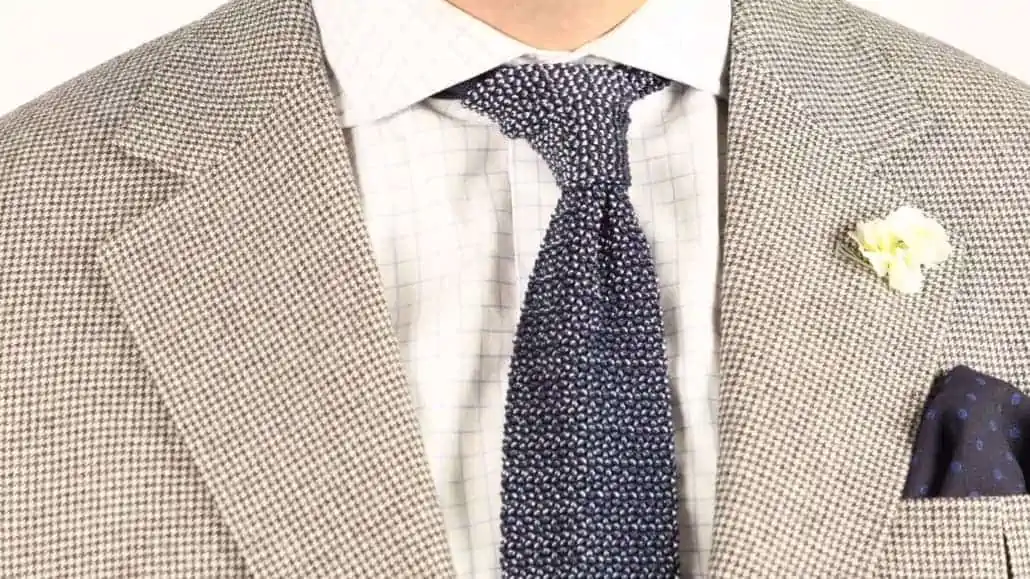 A navy blue wool challis pocket square with blue polka dots paired with a houndstooth jacket, a checked shirt, a white carnation boutonniere, and a navy and light blue two-tone knit tie.