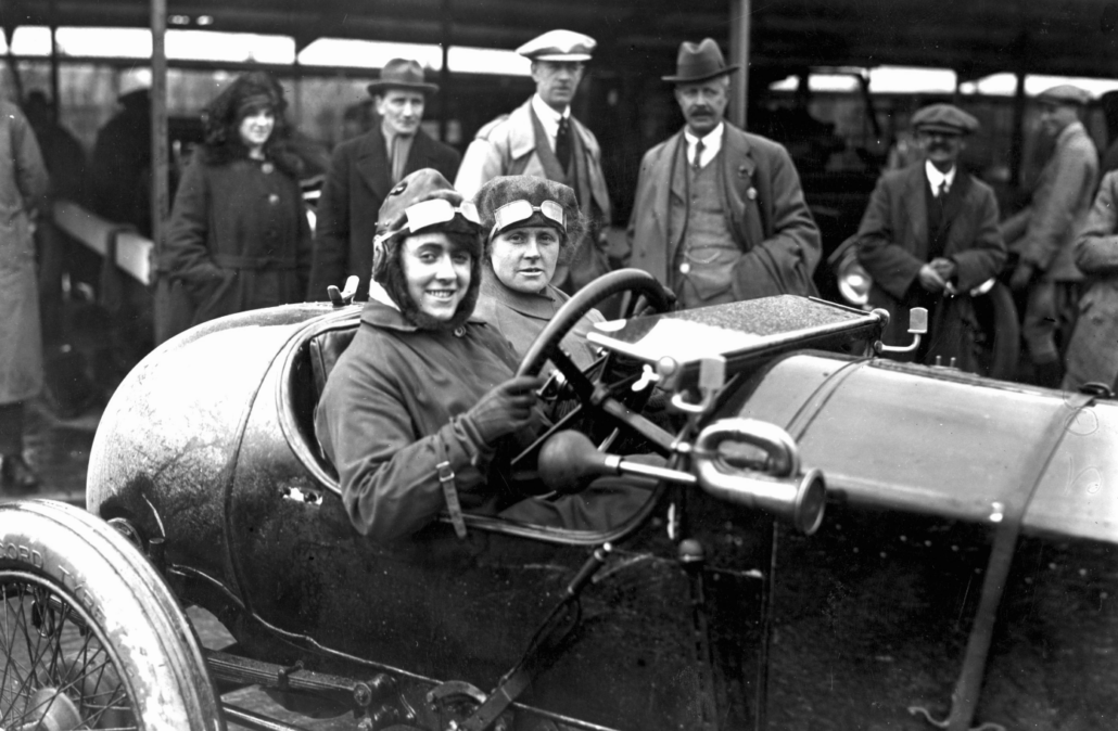 An illustration of a group of spectators admiring a 1920s style racing car. The men wear gloves.