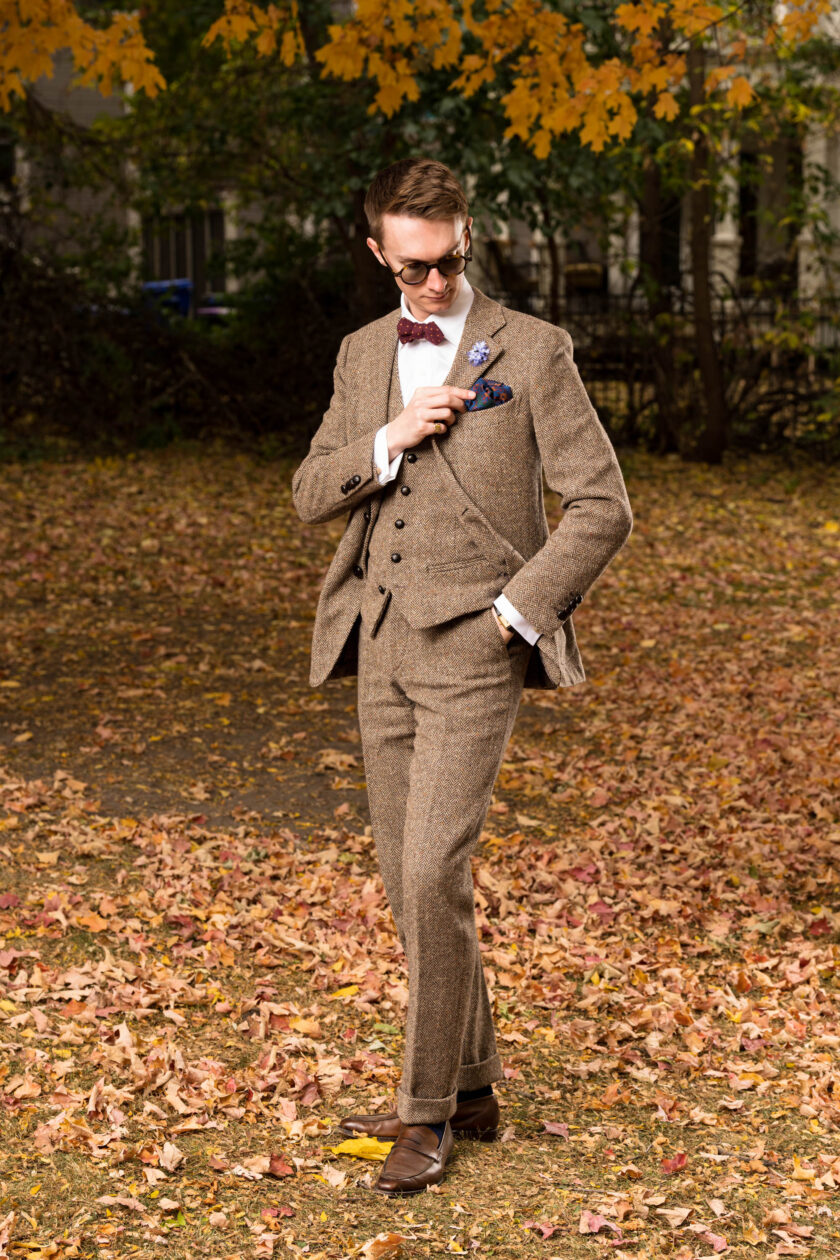 A photograph of a man wearing a light brown three piece suit in a park filled with leaves 