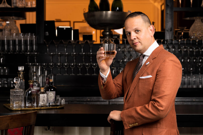 Raphael stands at a bar wearing a rust orange suit 