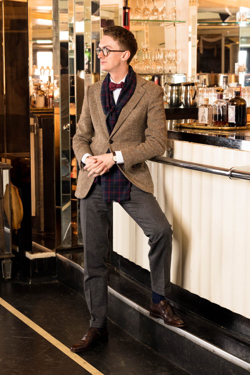 A man leans against a bar wearing a tan suit, grey trousers, and a plaid scarf with a red and green bowtie.  