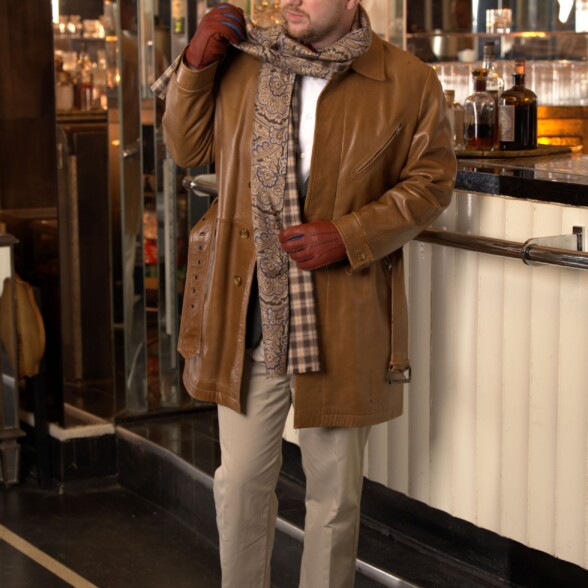 A man stands by a bar wearing a leather coat with dangling scarf ends
