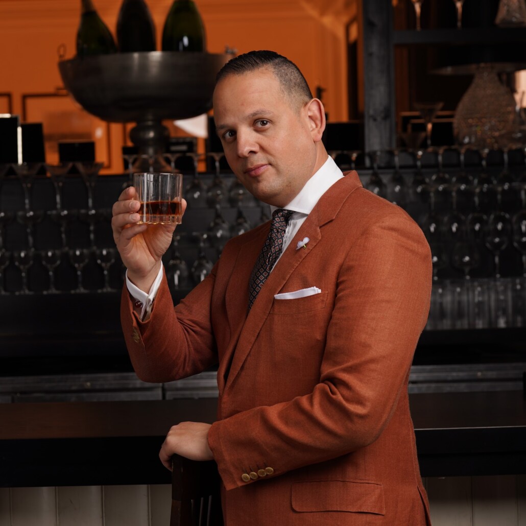Raphael stands at a bar wearing a rust orange suit holding a glass
