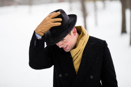 Raphael holds his hat while walking in the snow wearing a dark overcoat and yellow accessories